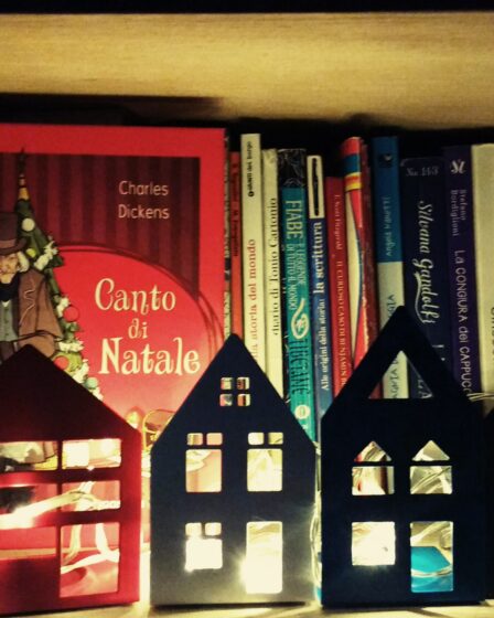 libri in uno scaffale libreria illuminato da case di Natale con lucine. In prima vista l'immagine del libro un Canto di Natale