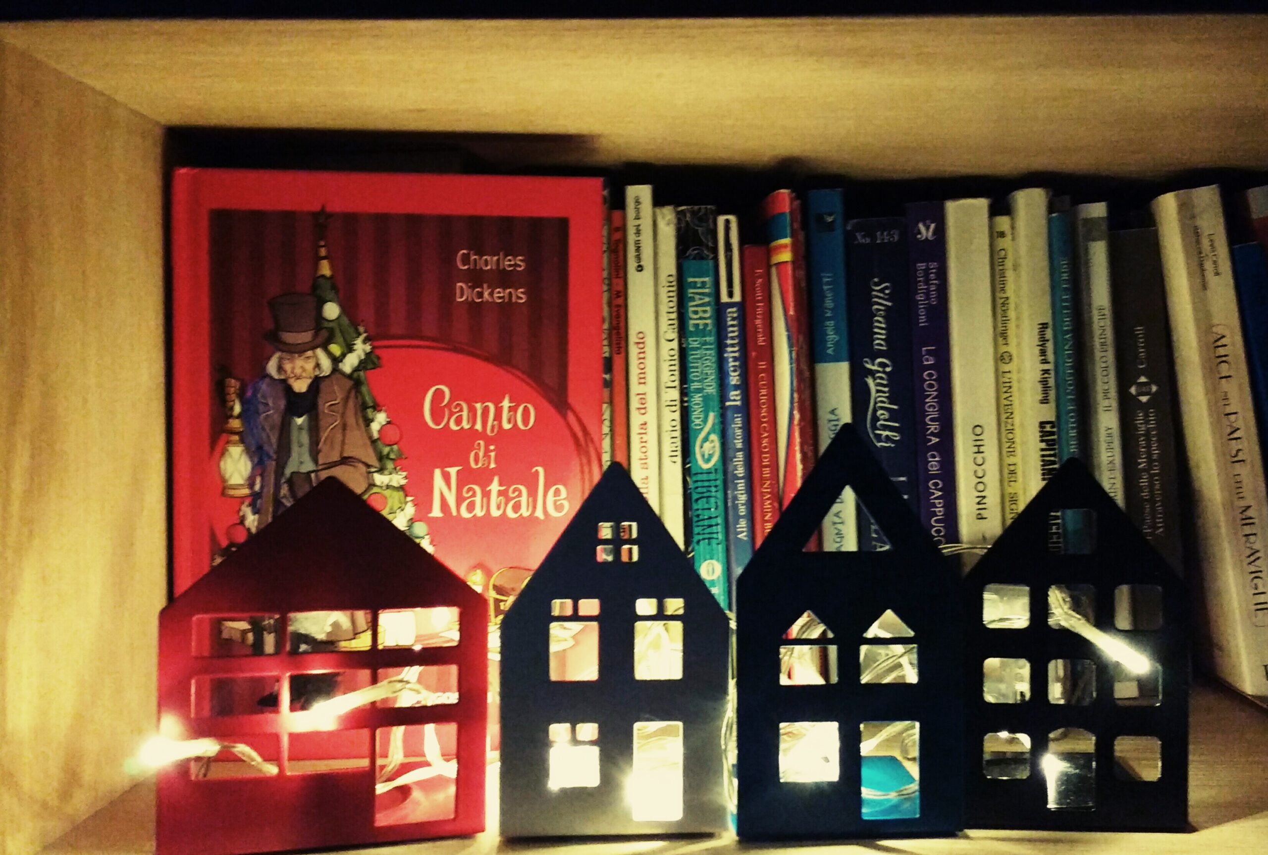 libri in uno scaffale libreria illuminato da case di Natale con lucine. In prima vista l'immagine del libro un Canto di Natale